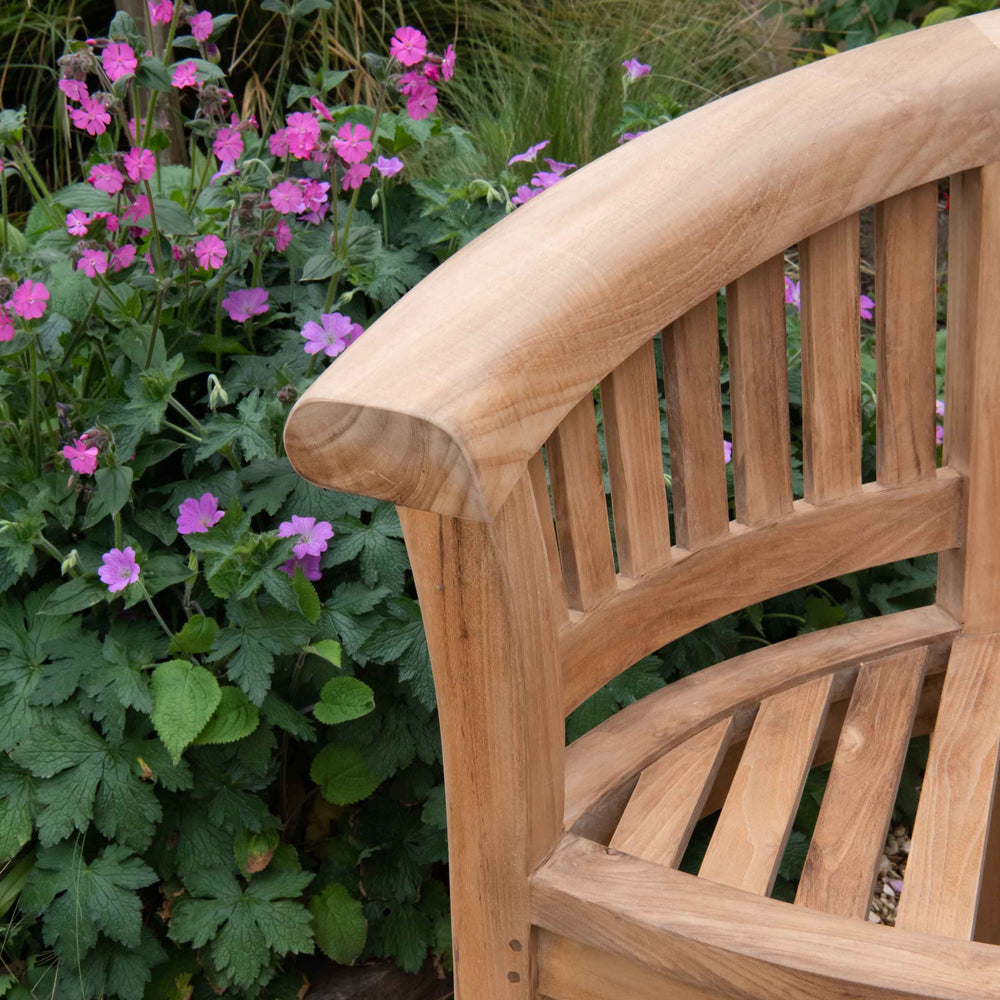 
                  
                    Close up of end of teak crescent bench with slatted seat and backrest and curved arms. The bench is in front of pink flowers in a flower bed. 
                  
                