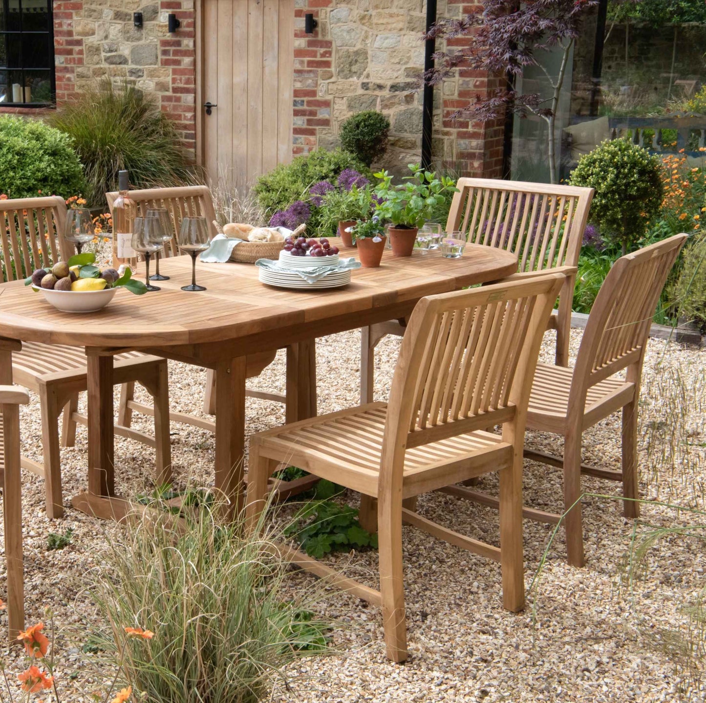 Teak oval dining table with teak dining chairs set around. The table holds bowl of fruit, wine glasses, plates and plants in pots and sits on gravel patio with flowerbeds behind. 